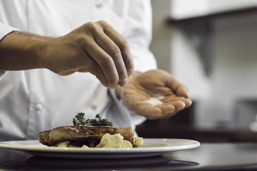 A chef's hand sprinkles salt over a dish, which features meat garnished with parsley and accompanied by vegetables, in a professional kitchen setting.