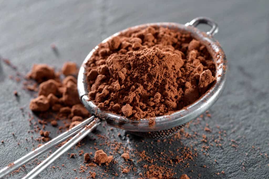 A metal sieve filled with cocoa powder rests on a dark surface, with some powder spilling out around it.