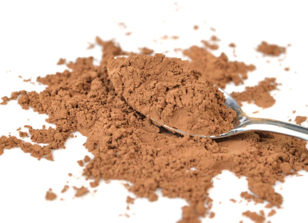 A silver spoon holding a pile of brown cocoa powder, with additional cocoa powder scattered on the white surface around it.