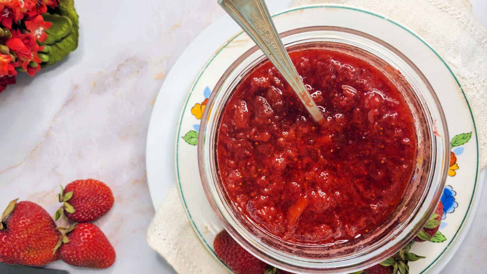 A jar of strawberry jam with a spoon inside, placed on a floral plate with a white cloth under it. Fresh strawberries are nearby on the left.