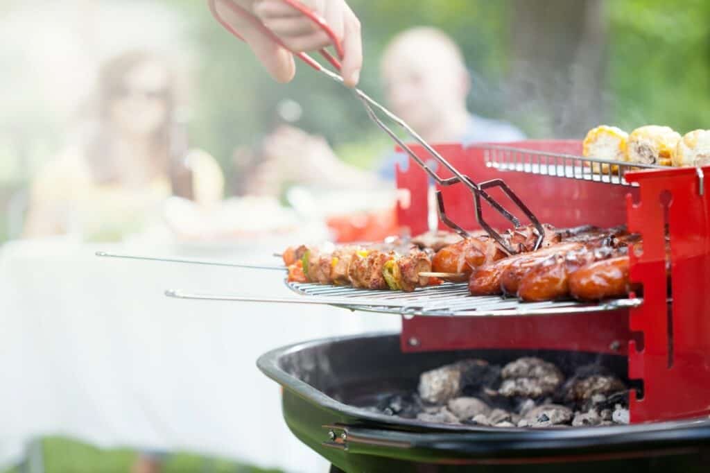 Close-up of a person grilling skewered vegetables and sausages on a red barbecue grill. Two people are blurred in the background sitting at a table with food. The scene is outdoors.