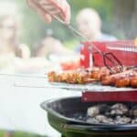 Close-up of a person grilling skewered vegetables and sausages on a red barbecue grill. Two people are blurred in the background sitting at a table with food. The scene is outdoors.