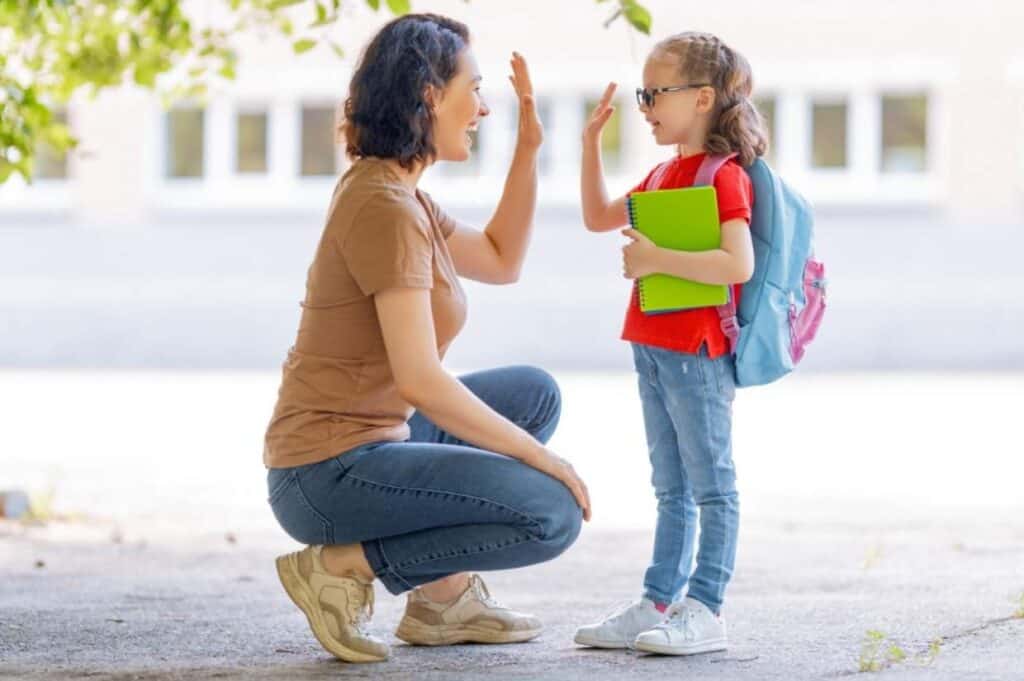 An adult woman kneels to high-five a young girl wearing a backpack and holding a notebook, both smiling outdoors near a building, celebrating back to school success.