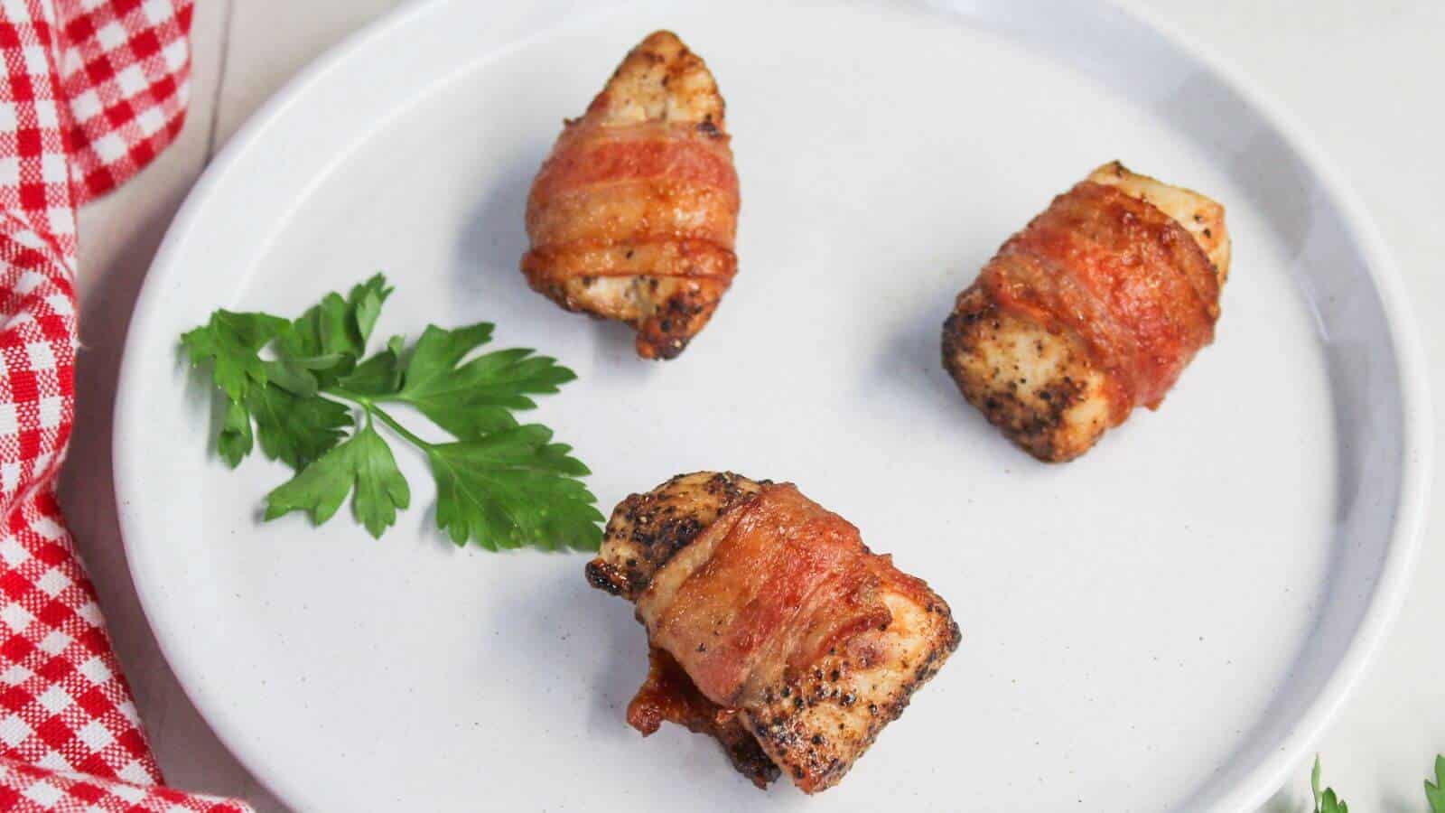 Three bacon-wrapped pieces of chicken on a white plate with a sprig of parsley and a red and white checkered cloth on the side.