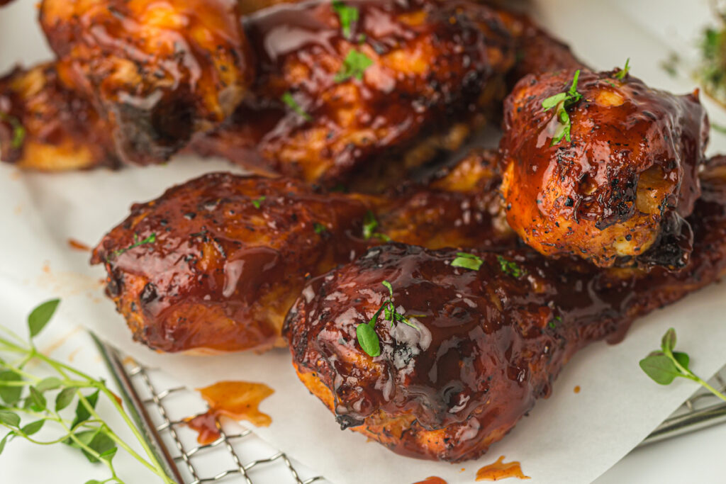 Barbecued chicken drumsticks coated in a thick, glossy sauce, garnished with fresh herbs, served on parchment paper on a wire rack.