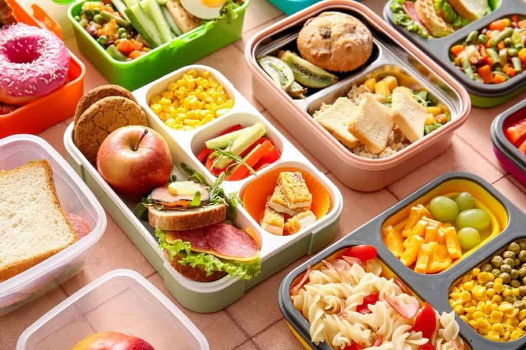 An assortment of colorful bento lunch boxes filled with various foods including sandwiches, fruits, vegetables, pasta, a donut, and other snacks, arranged neatly on a table.
