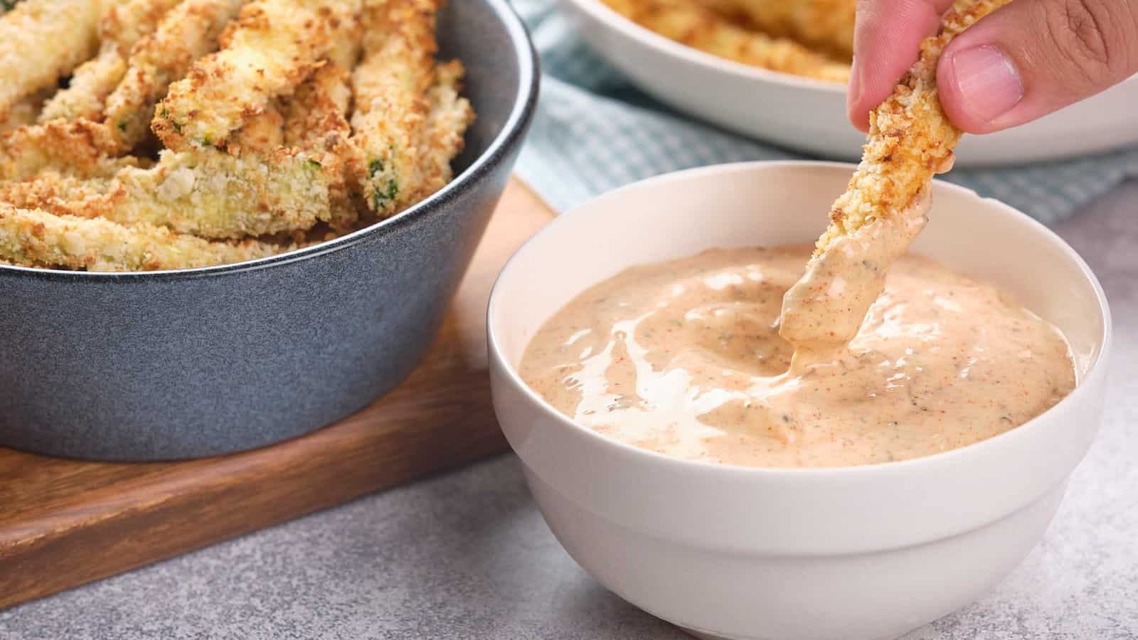 A hand dips a crispy breaded zucchini stick into a bowl of blackened ranch sauce.