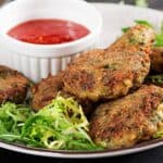 A plate of canned salmon burgers on a bed of greens, served with a side of red dipping sauce in a small white bowl.