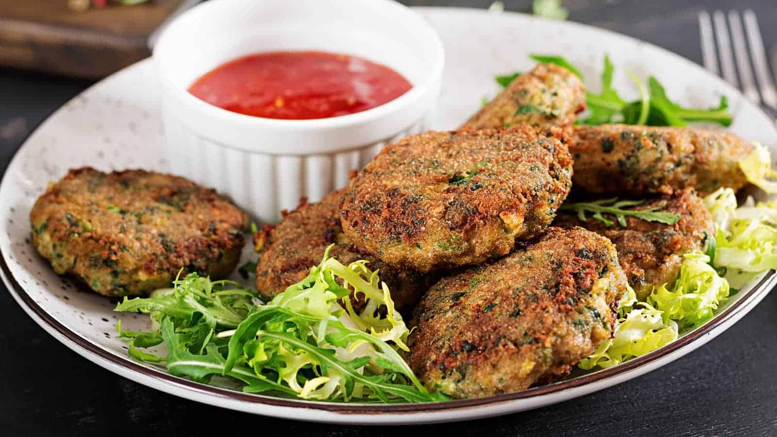 A plate of canned salmon burgers on a bed of greens, served with a side of red dipping sauce in a small white bowl.