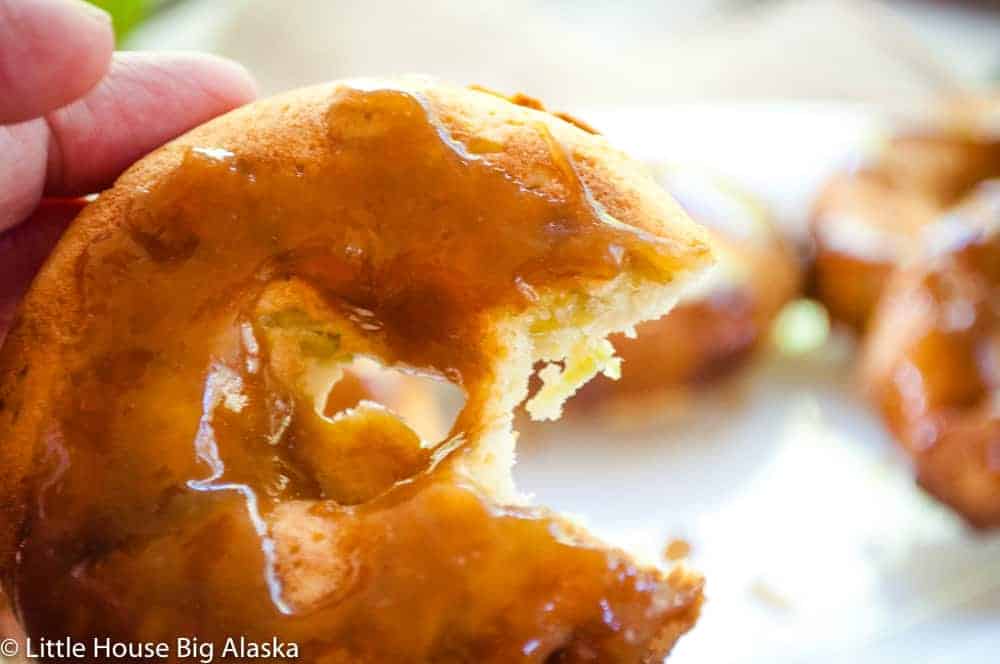 A person holds a caramel-glazed donut with a bite taken out. More donuts are blurred in the background.