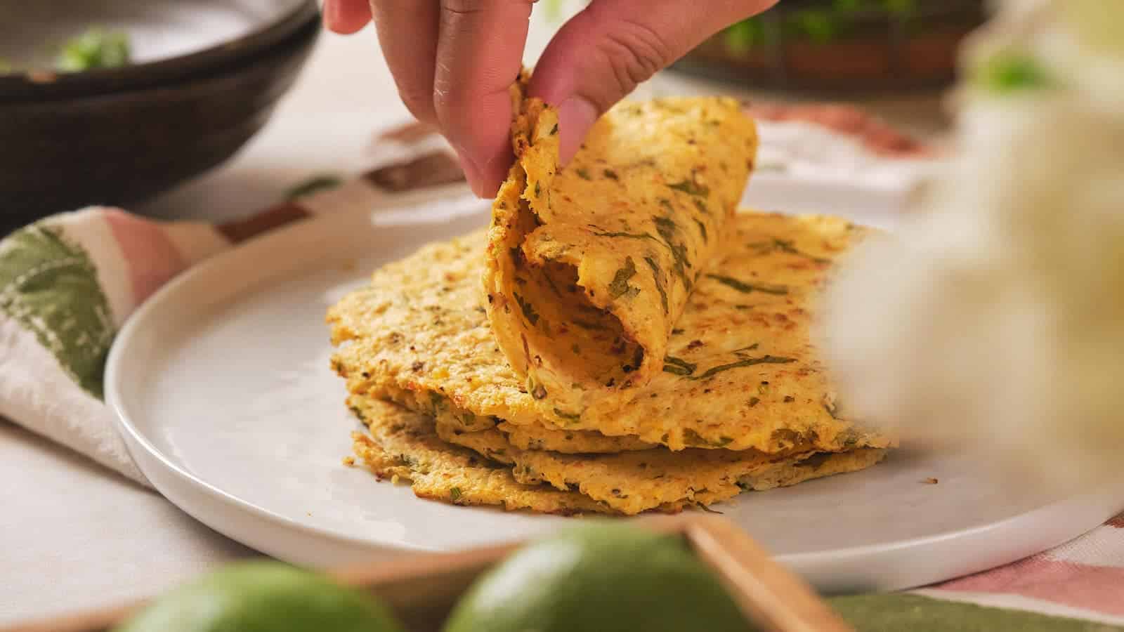 A hand holds and begins to wrap a folded cauliflower tortilla, which is on a white plate.