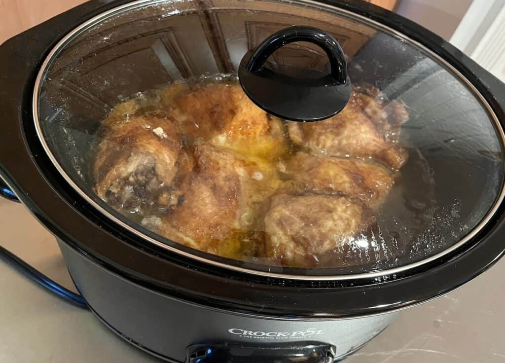 A slow cooker filled with cooking chicken thighs, partially submerged in liquid, with a glass lid on.