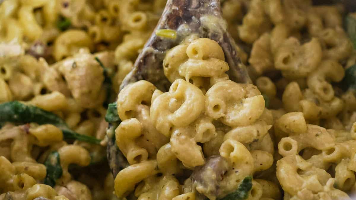 Close-up of creamy macaroni and cheese with visible greens and mushrooms, being scooped with a wooden spoon.