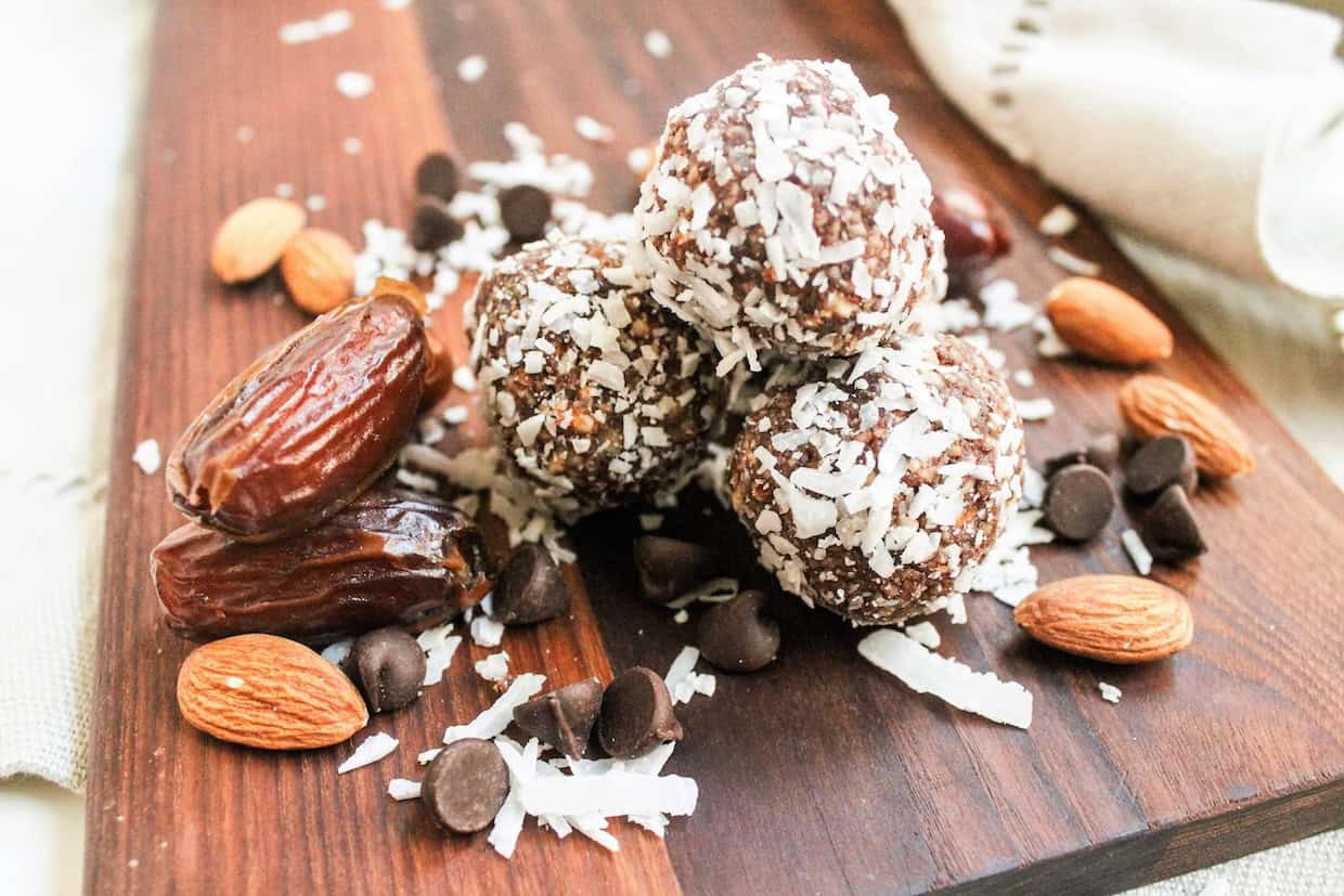 A wooden board with energy balls coated in shredded coconut, surrounded by dates, almonds, and chocolate chips.