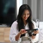 A person with long dark hair sits on a couch, smiling while playing a cozy game that is not a farming sim on a handheld console controller. A TV and dresser are in the background.