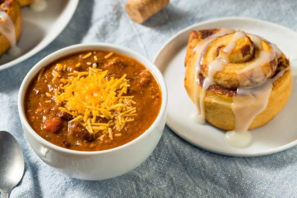 A bowl of chili topped with shredded cheese is next to a cinnamon roll with icing on a white plate.