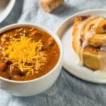 A bowl of chili topped with shredded cheese is next to a cinnamon roll with icing on a white plate.