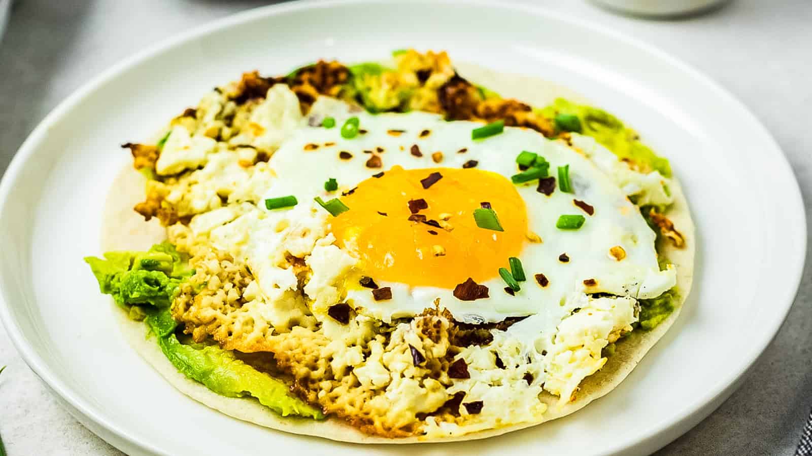 A fried egg with crispy feta cheese, avocado, and a tortilla, garnished with chopped green onions and red pepper flakes, served on a white plate.