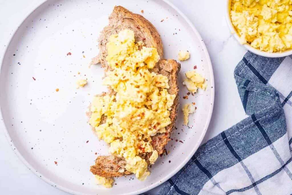 A white plate with a piece of bread topped with scrambled eggs. A small bowl of additional scrambled eggs and a blue and white checkered cloth napkin are beside the plate.