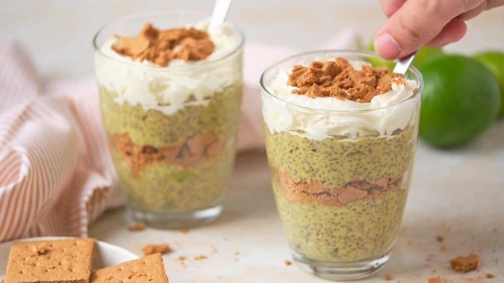 Two glasses of chia seed pudding layered with crushed graham crackers and topped with whipped cream, perfect for delightful breakfasts. A hand holds a spoon in one glass, ready to indulge. A striped cloth and limes add a fresh backdrop.