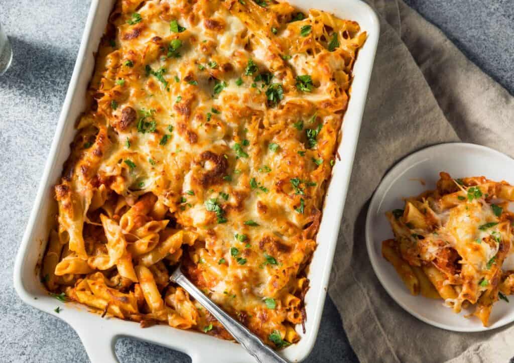 A baked pasta dish with melted cheese and garnished with chopped herbs in a white rectangular dish. A serving of the pasta is placed on a white plate beside it, with a napkin underneath.