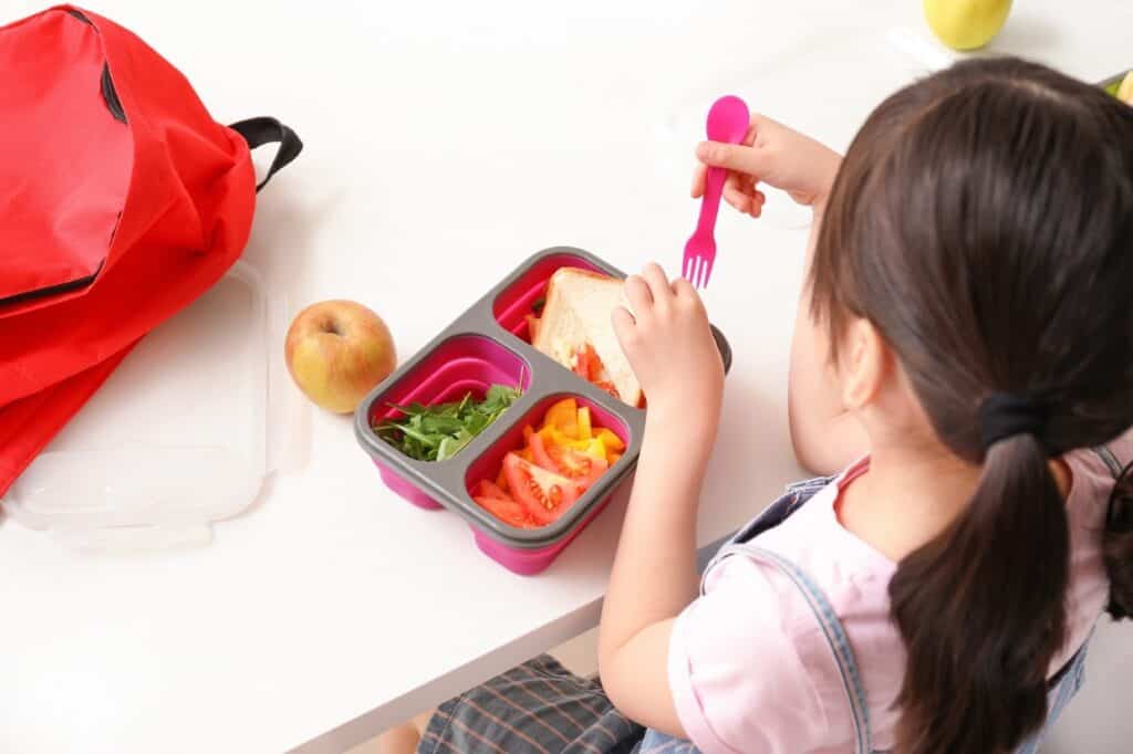 A child with brown hair sits at a table eating from a colorful bento lunch box filled with a sandwich, sliced tomatoes, bell peppers, and greens. A red bag and an apple are on the table.