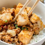 A bowl of rice topped with stir-fried tofu cubes and minced vegetables, garnished with green onions. Chopsticks rest on top.