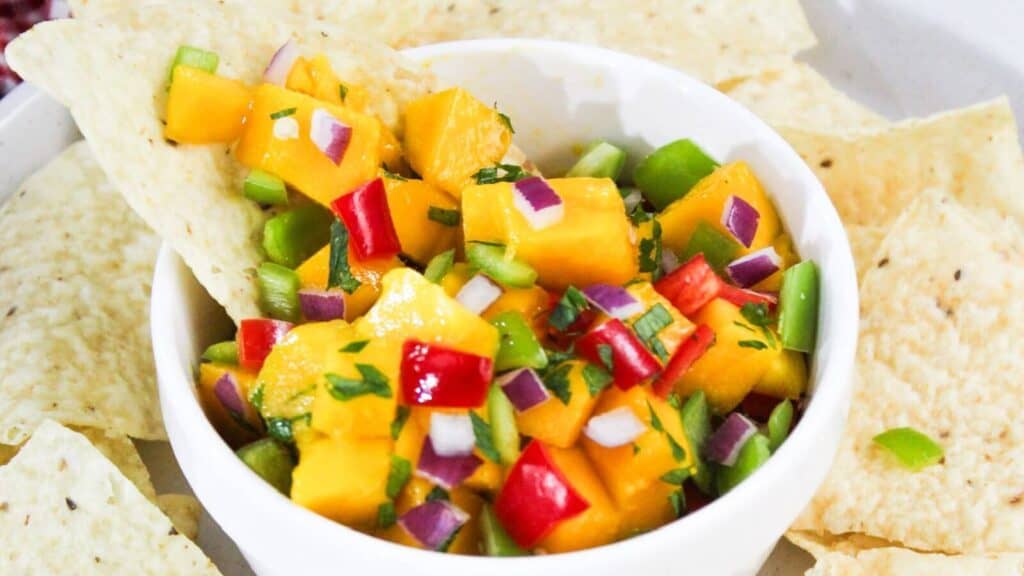 A bowl of mango salsa with diced mango, red onions, red and green bell peppers, and cilantro, surrounded by tortilla chips.