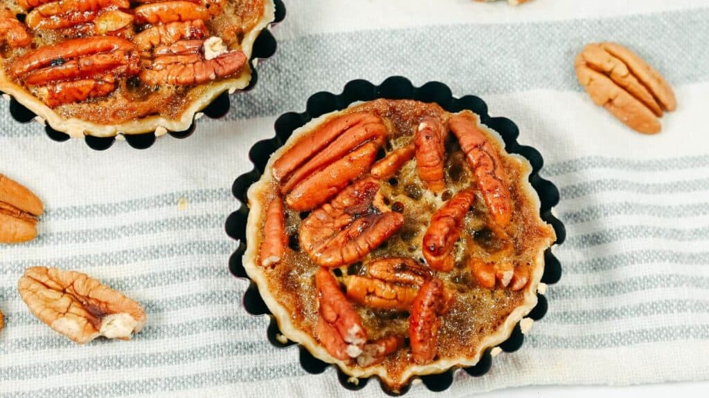 Two pecan tarts on a striped cloth, surrounded by pecan halves.