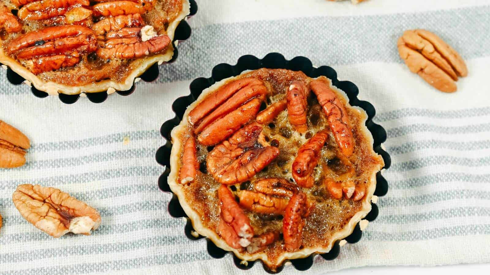 Two pecan pie tarts placed on a striped cloth with scattered pecans around them.