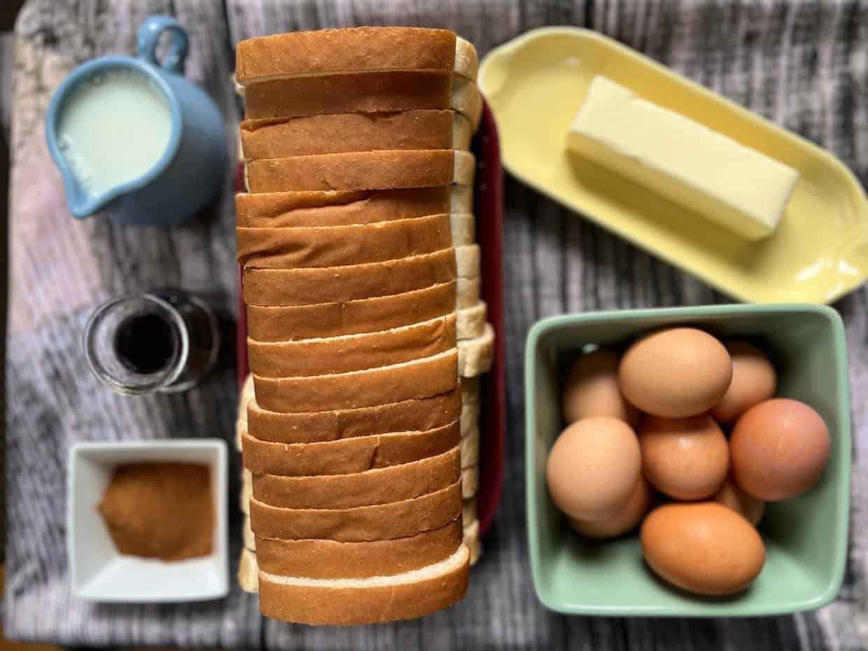 A loaf of sliced bread, a butter dish with a stick of butter, a bowl of eggs, a small jug of milk, a small jar of jam, and a dish of brown sugar arranged on a table.