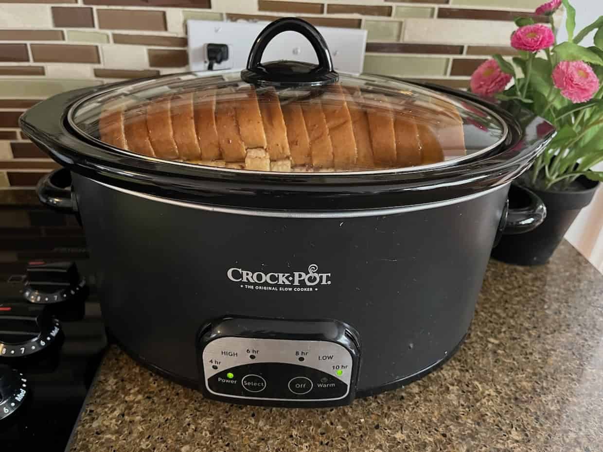 A Crock-Pot slow cooker on a kitchen counter contains a loaf of sliced bread. A potted plant with pink flowers and a stovetop are visible in the background.