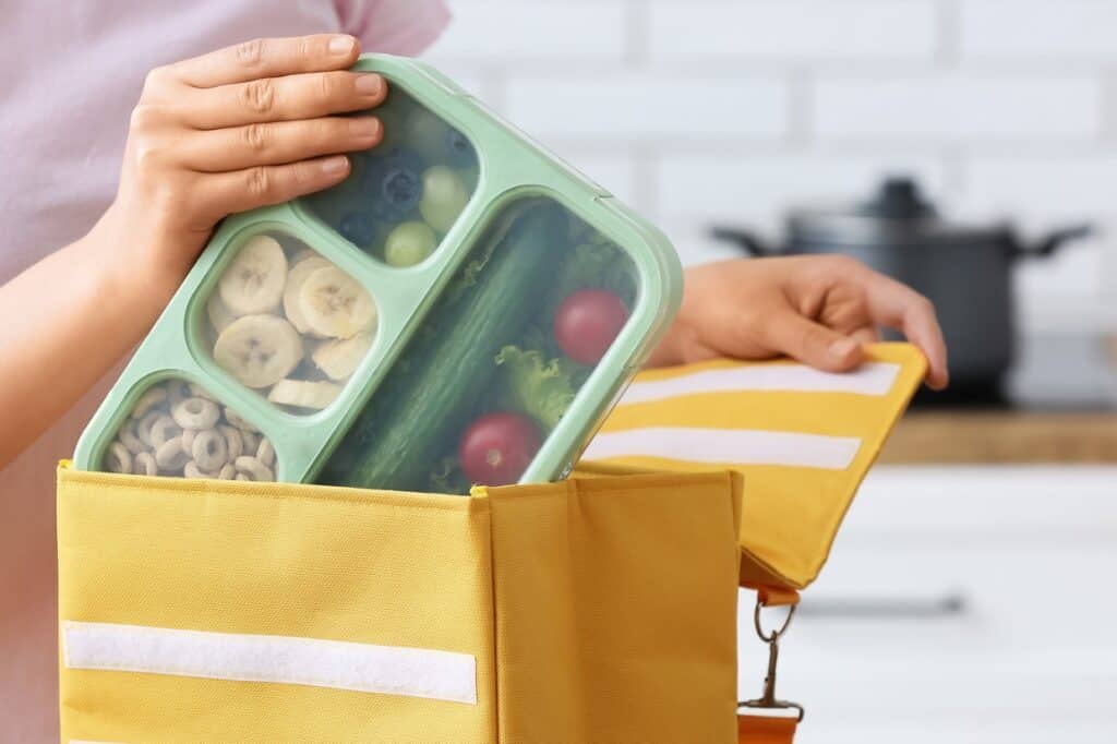 Person carefully placing a green bento lunch box filled with compartments of fruits, vegetables, and cereal into a yellow insulated lunch bag in a kitchen.
