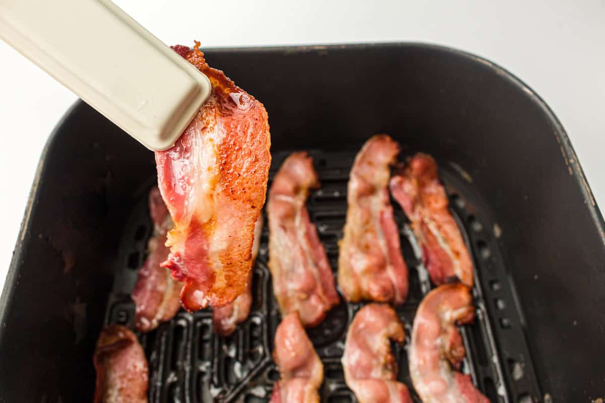 Cooking bacon strips in an air fryer. One strip held by tongs is shown above other strips inside the air fryer basket.