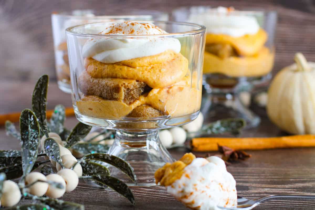 Three glass dessert cups filled with layers of pumpkin mousse, whipped cream, and sponge cake sit on a wooden surface, adorned with greenery and cinnamon sticks. A small white pumpkin is in the background.