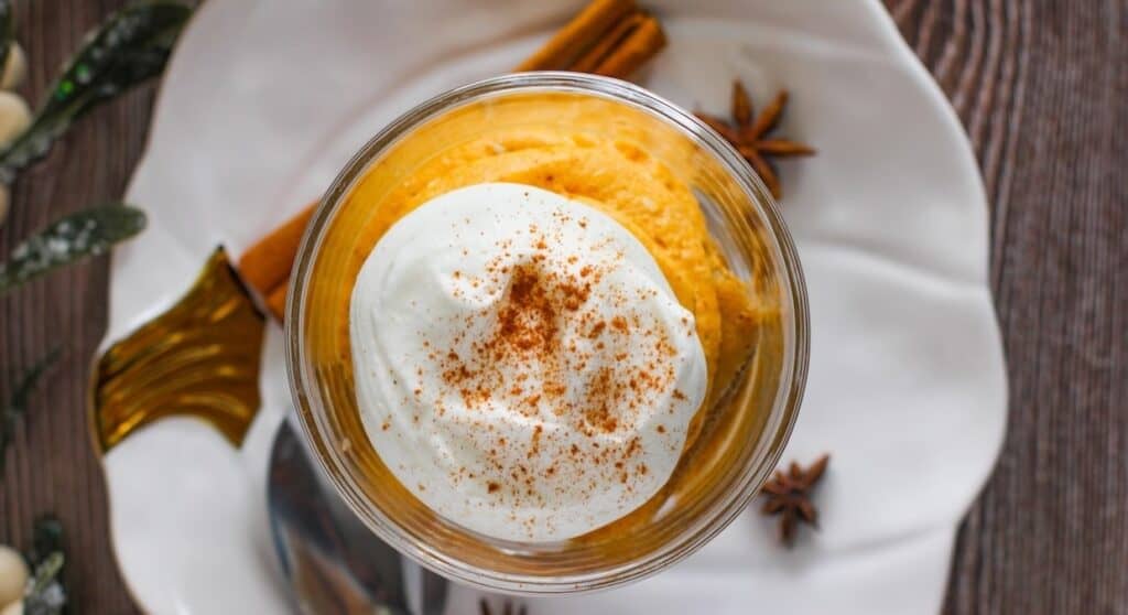 A dessert glass filled with pumpkin mousse, topped with whipped cream and cinnamon, is placed on a white plate with a cinnamon stick and star anise ornaments.