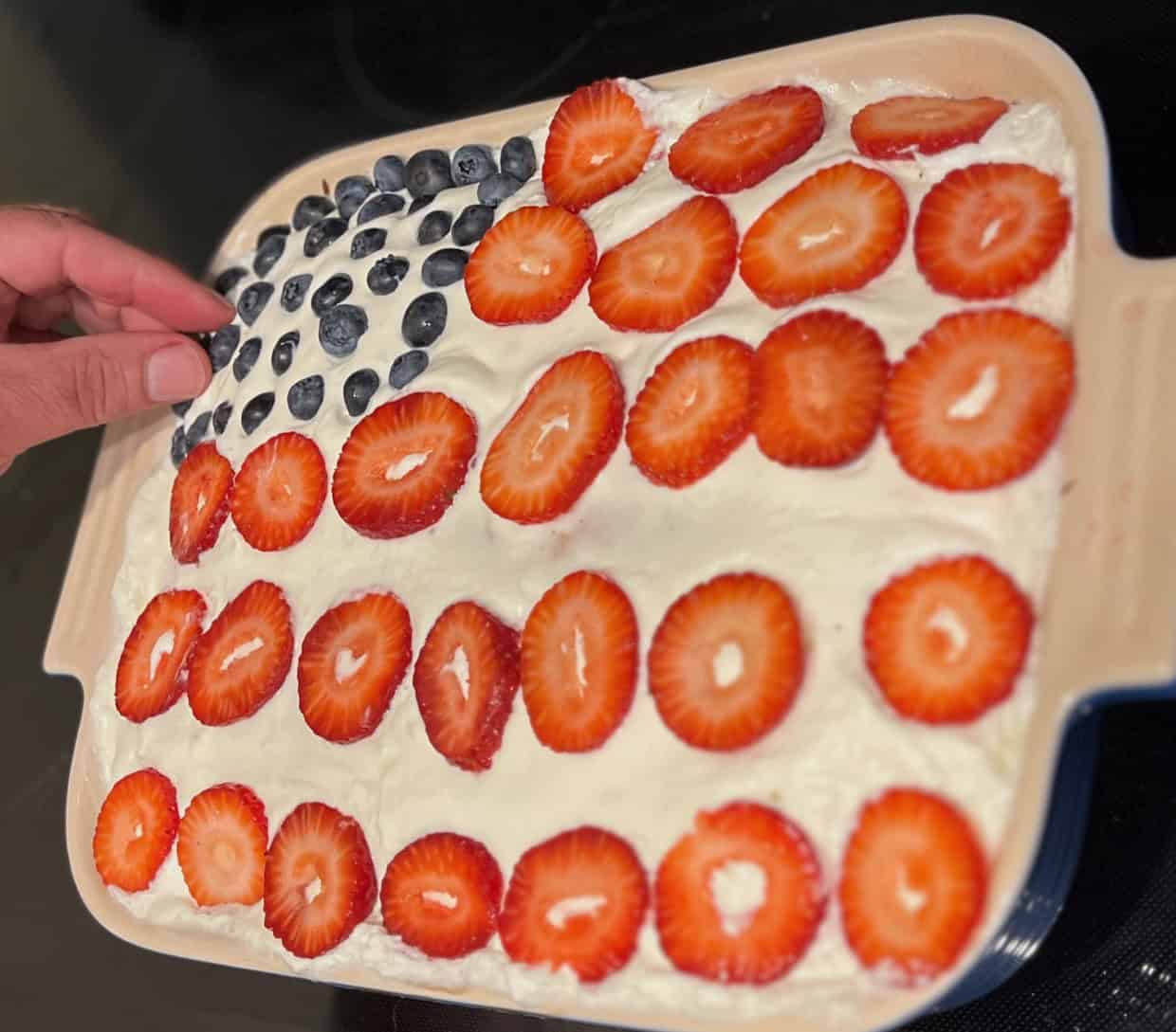 A dessert topped with blueberries in the shape of a corner rectangle and strawberry slices arranged to resemble the American flag. A hand is adding a blueberry.