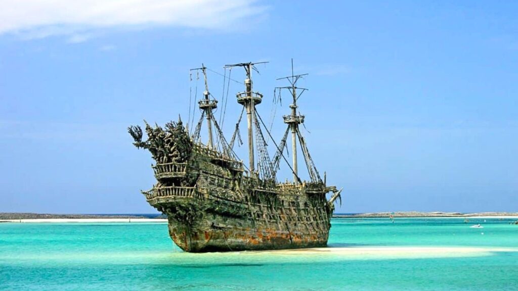 A large, weathered, and ornate pirate ship with tall masts floats in shallow turquoise water near a sandy shoreline under a clear sky, evoking the vibrant history of Nassau and its legendary era of swashbuckling pirates.