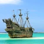 A large, weathered, and ornate pirate ship with tall masts floats in shallow turquoise water near a sandy shoreline under a clear sky, evoking the vibrant history of Nassau and its legendary era of swashbuckling pirates.