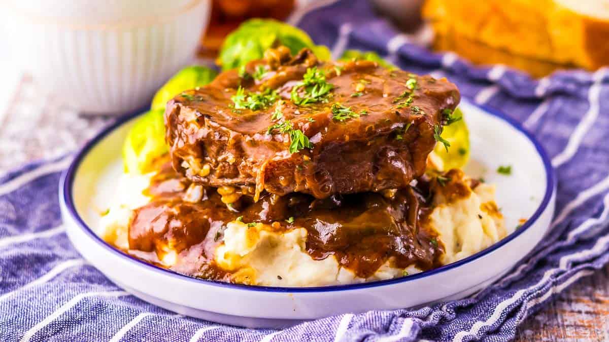 A plate with a serving of mashed potatoes topped with two pieces of meat in brown gravy, garnished with herbs and accompanied by Brussels sprouts.