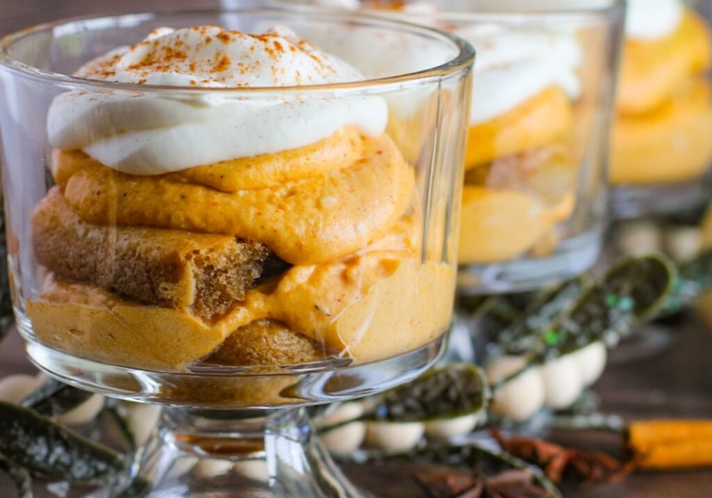 A close-up view of a dessert in a glass cup featuring layers of creamy pumpkin custard and whipped topping, with additional cups in the background and decorative foliage around.