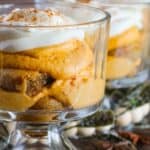 A close-up view of a dessert in a glass cup featuring layers of creamy pumpkin custard and whipped topping, with additional cups in the background and decorative foliage around.
