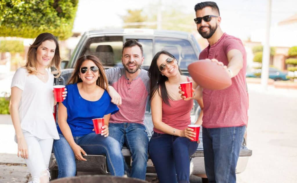 Five people smiling, sitting on the tailgate of a truck, holding red cups. One person is holding a football and wearing sunglasses. They appear to be enjoying an outdoor casual gathering, embodying classic tailgating traditions.