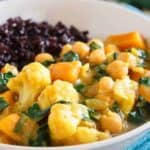 A white bowl containing a vegetable curry with cauliflower, chickpeas, and greens, served alongside a portion of dark-colored rice.