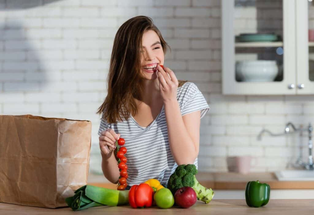 A woman stands in a kitchen, smiling while holding and eating cherry tomatoes from a bunch. Various vegetables and a paper grocery bag are on the counter in front of her.