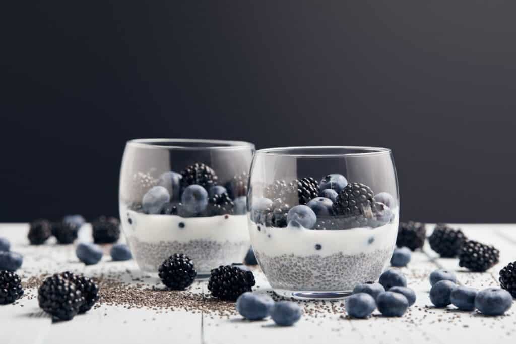 Two glass cups filled with chia pudding, layered with white yogurt and topped with blueberries and blackberries, on a surface scattered with extra berries and chia seeds.