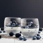 Two glass cups filled with chia pudding, layered with white yogurt and topped with blueberries and blackberries, on a surface scattered with extra berries and chia seeds.