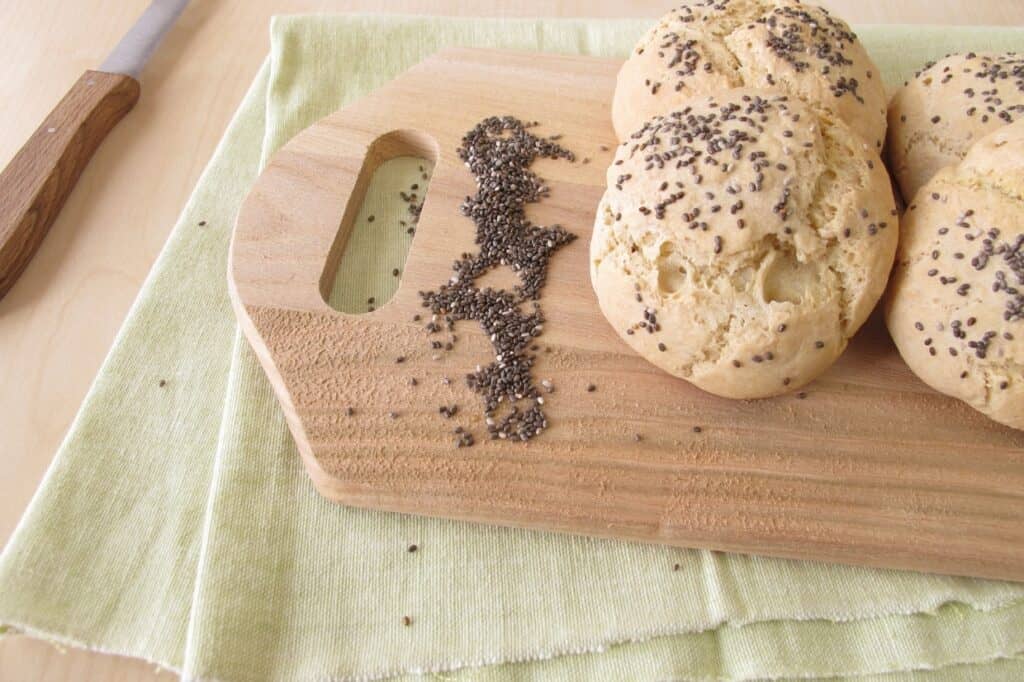 Three bread rolls with chia seeds on a wooden cutting board next to a scattering of chia seeds on a green cloth, with a knife in the background.