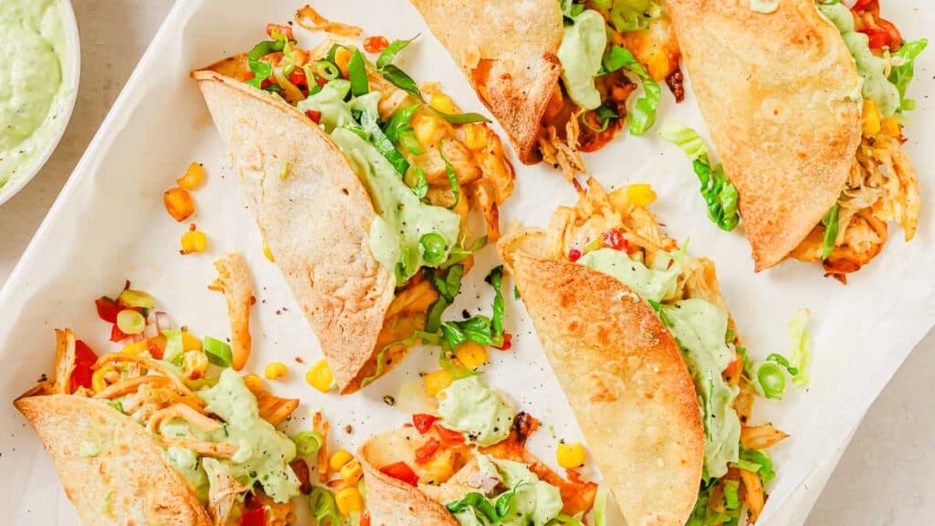 A tray of crispy tacos filled with shredded chicken, corn, bell peppers, scallions, and topped with a creamy green sauce.