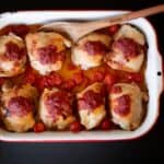 A baking dish contains eight baked chicken pieces topped with tomato sauce and surrounded by roasted cherry tomatoes, with a wooden spoon resting on the edge.